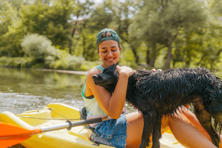Photo of a young woman kayaking down the river and enjoying beautiful nature that surrounds it. She has a companion - her furry best friend - her pet dog.